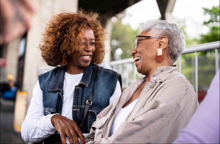 Photo of a African-American woman talking to an older African-American woman sitting outside, both with COVID-19 high risk factors.